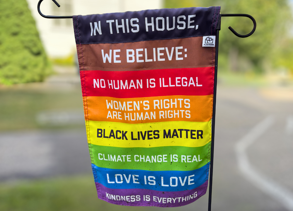 Flag outside reads, "In this House, We Believe No human is illegal, Women's rights are human rights, Black Lives matter, Climate change is real, Love is Love, Kindness is Everything
